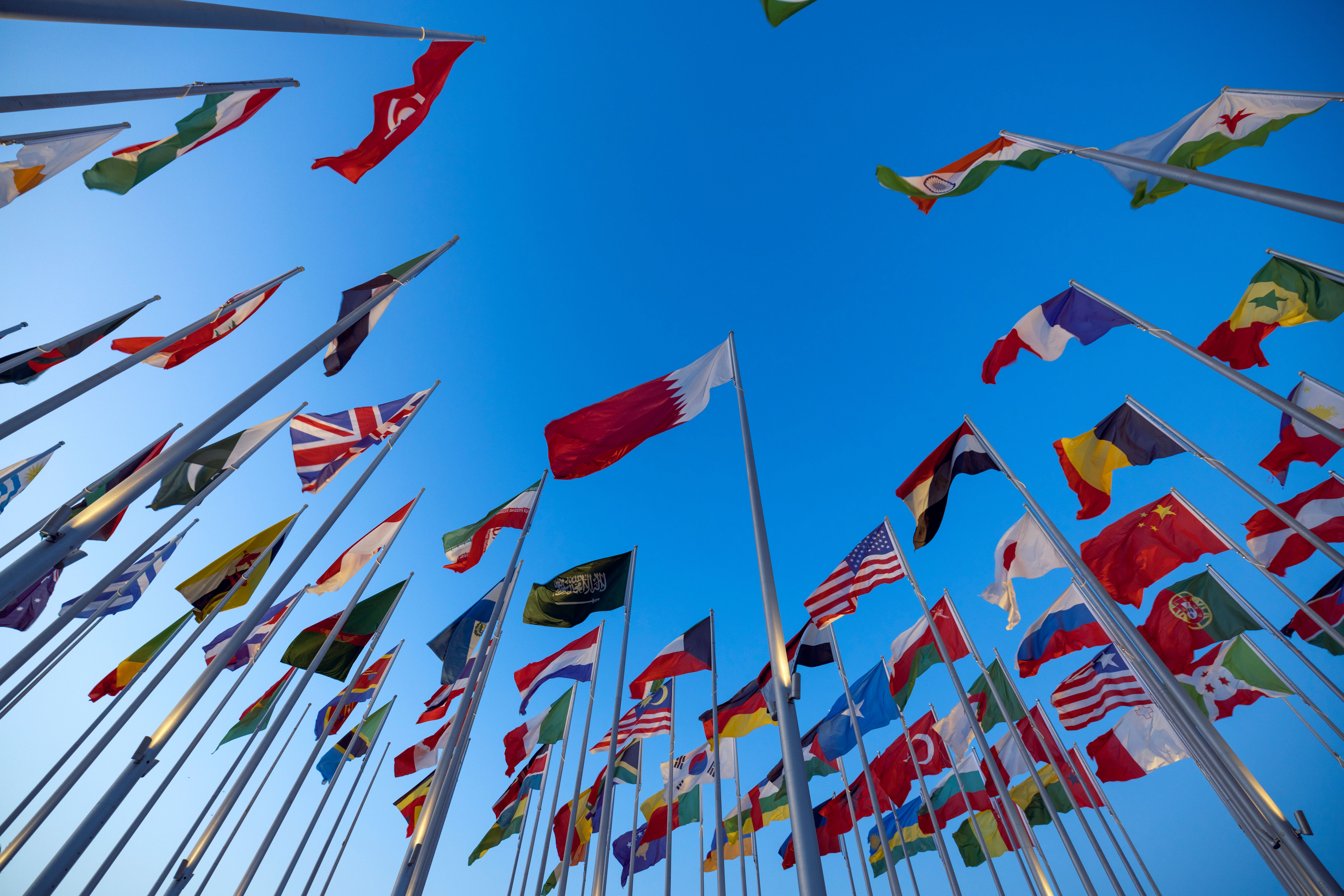 Credit: Pexels License, via https://www.pexels.com/photo/low-angle-shot-of-flags-on-the-background-of-a-clear-blue-sky-14676984/ Dozens of national flags on tall flagpoles in a circular formation against a clear blue sky