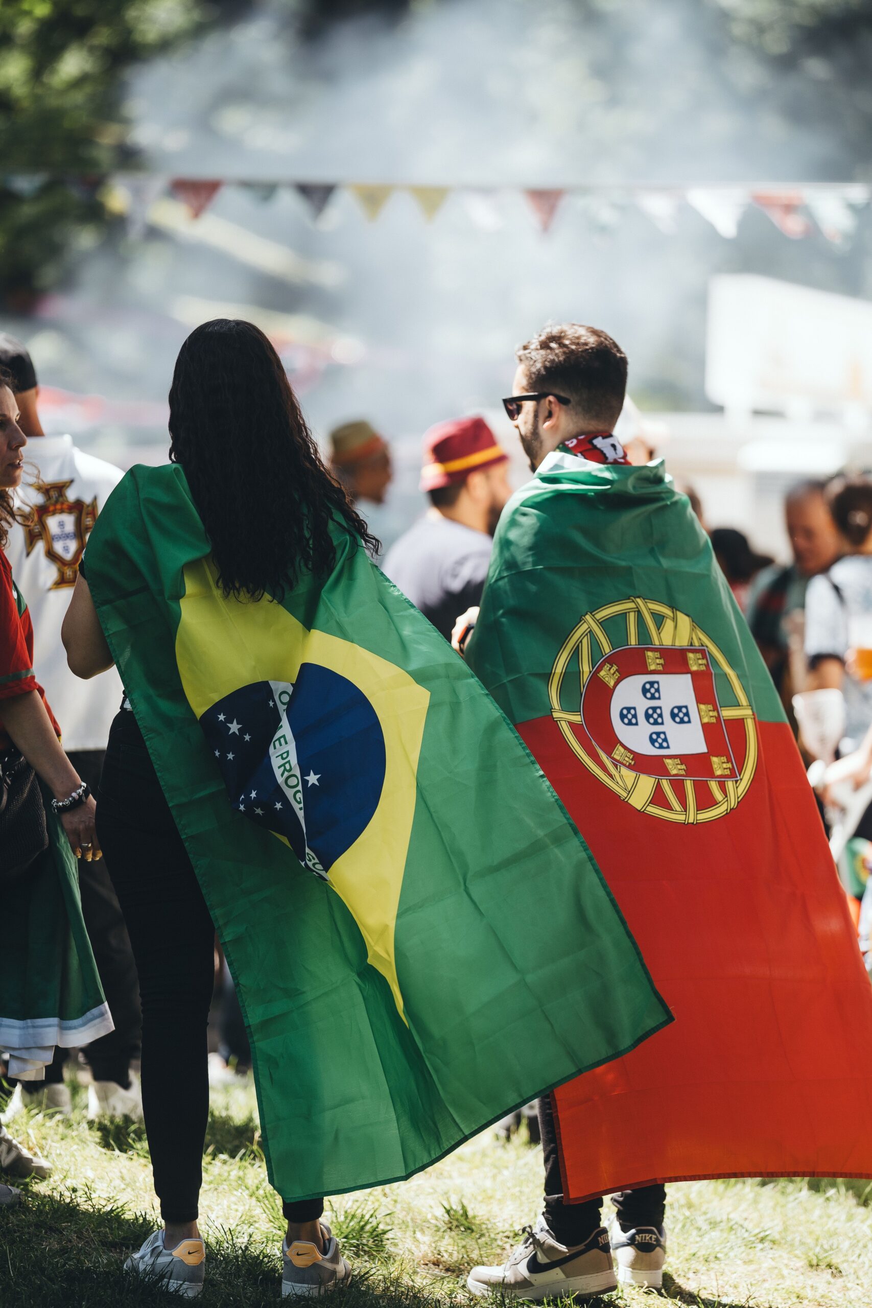 Two people at an outdoor gathering wearing Brazilian and Portuguese flags over their shoulders