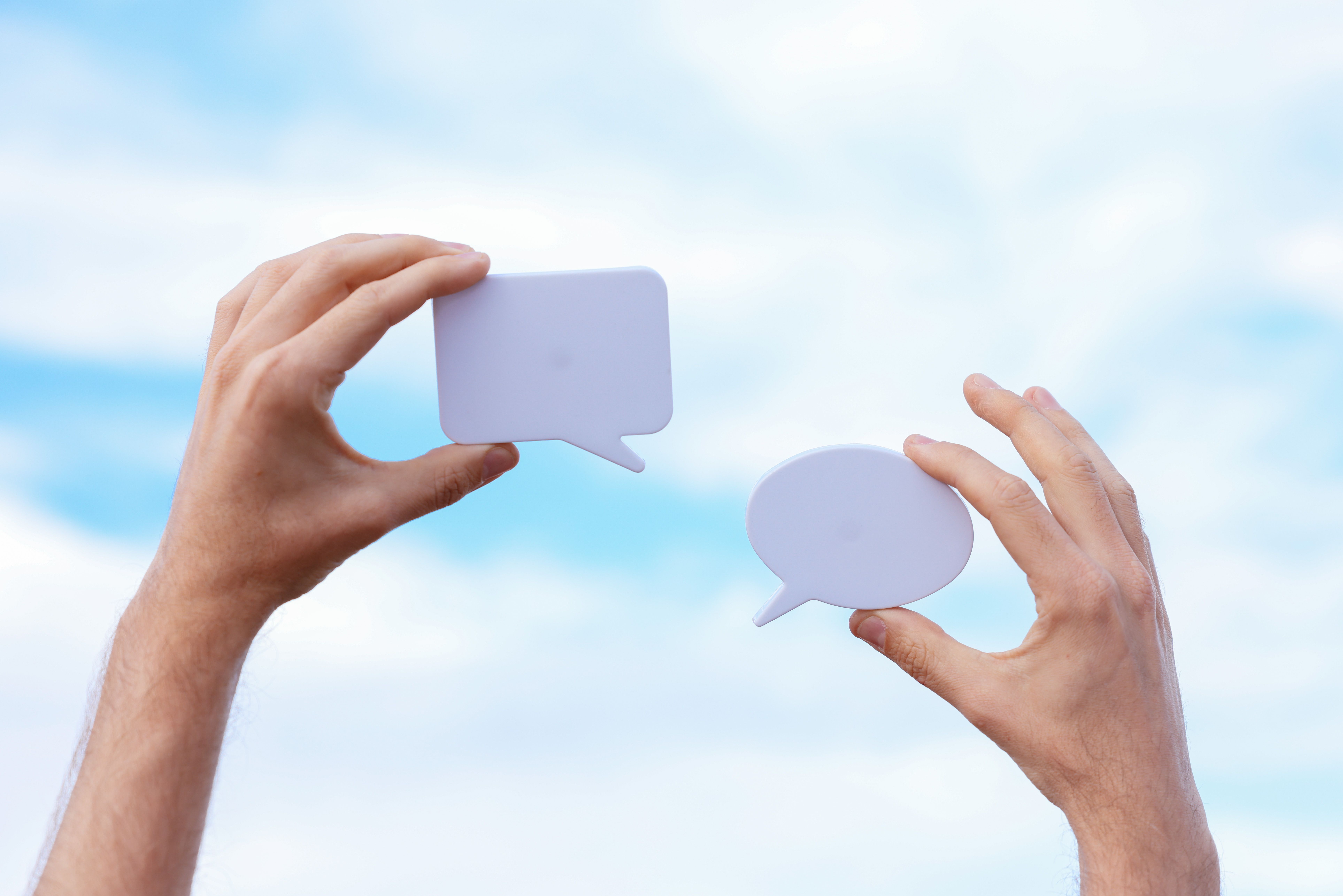 Credit: cup-of-couple, Pexels License, via https://www.pexels.com/photo/close-up-of-man-holding-paper-cutouts-in-the-shape-of-conversational-clouds-8015694/ Two hands holding blank speech-bubble cutouts against a sky, representing two people in dialogue